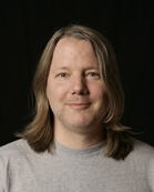 A man with shoulder-length light brown hair and a grey t-shirt smiling against a black background.