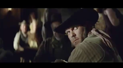 A man wearing a dark wide-brimmed hat looks back over his shoulder in a dark, crowded room.
