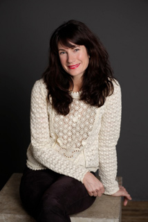 A woman with long dark hair and red lipstick wearing a white textured lace sweater, sitting against a dark background.