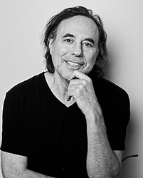 Black and white portrait of a smiling man with long hair resting his chin on his hand.
