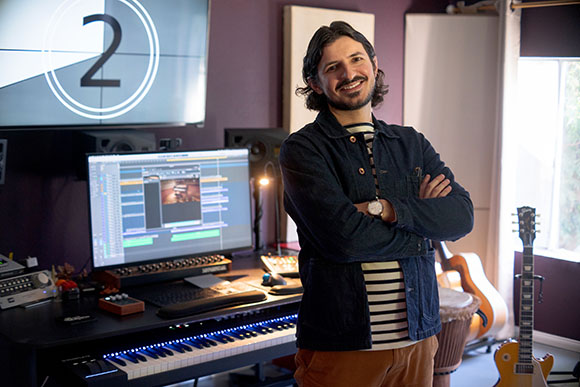 A smiling man with dark hair and a beard stands in a music studio with a computer, keyboard, and guitars.