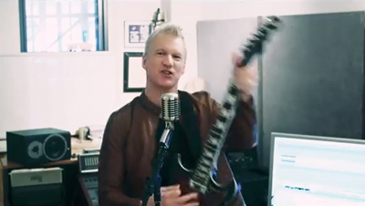 A man in a brown shirt holds an electric guitar in front of a microphone in a studio.