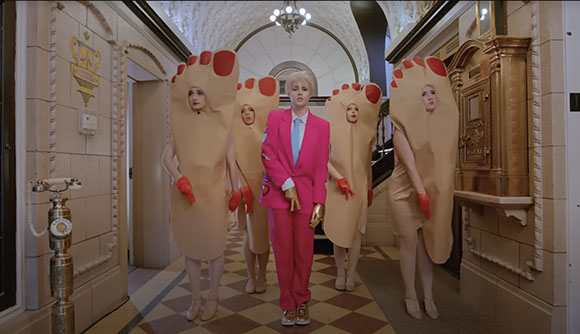 A person in a pink suit stands in a hallway flanked by four people wearing large tan foot costumes.