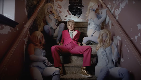 A man in a pink suit sits on stairs surrounded by four women in light blue lingerie.
