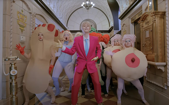 People in avant-garde costumes, including large beige shapes, pose in a grand, arched hallway.