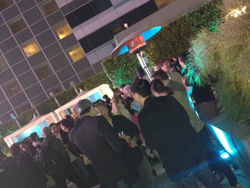 A crowd of people stand together outdoors at night, with building lights and greenery visible.