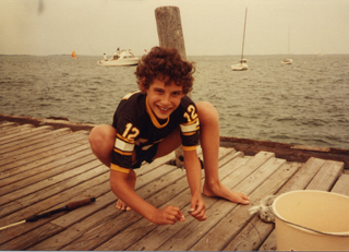 A young boy in a black and yellow jersey crouches on a wooden pier near a body of water.