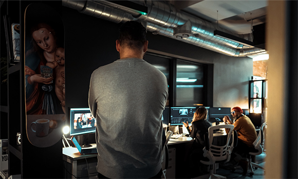 A man seen from behind in a dark office looking toward two people working at desks with multiple monitors.