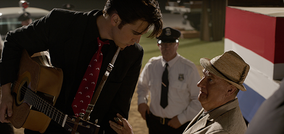 A young man in a black suit and red tie holds an acoustic guitar, looking down at an older man.