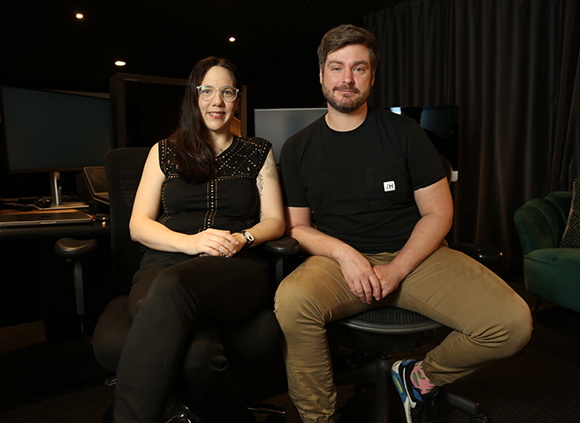 A woman in a black top and a man in a black t-shirt sit side-by-side in office chairs.