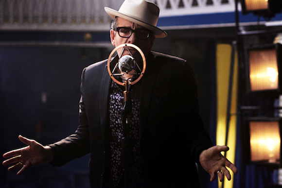 A man in a white fedora and glasses sings into a circular vintage microphone with his hands outstretched.