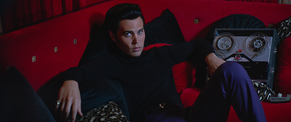 A man in a black turtleneck sits on a red velvet sofa next to a vintage reel-to-reel tape recorder.