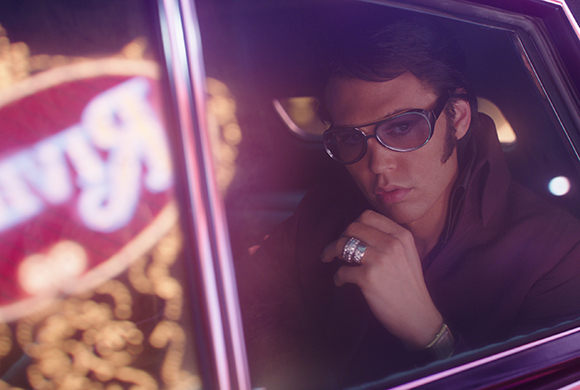 A man wearing dark sunglasses looks out from the backseat of a car at night.