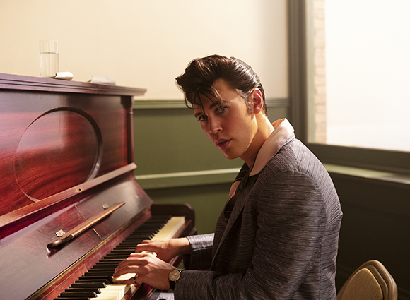 A man with dark pompadour hair sits at a dark wood piano, looking toward the camera.