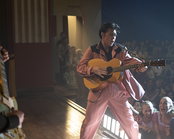 A man in a pink suit plays an acoustic guitar on a stage in front of a cheering crowd.
