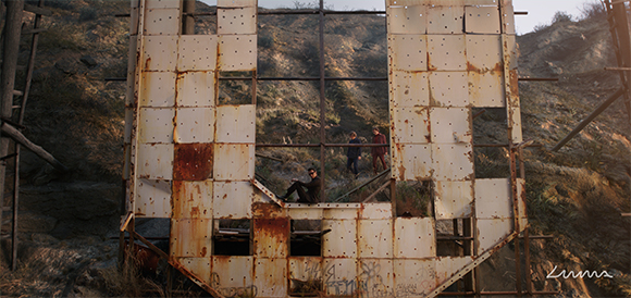 Two people stand behind a large, rusted metal structure shaped like a letter B in a hilly, brushy landscape.