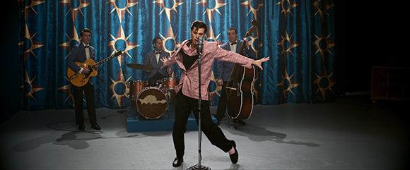 A singer in a pink jacket performs with a band against a blue curtain with gold star patterns.