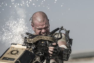 A man with a strained expression operates a large mechanical device as bright sparks fly around him.