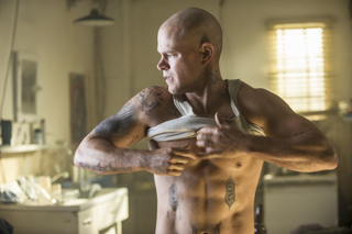 A muscular, bald man with tattoos pulls up his white tank top in a dimly lit room.
