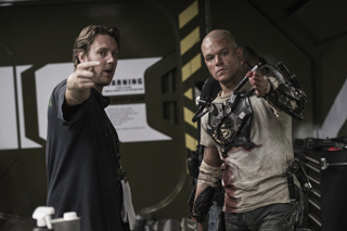 A man in a dark shirt points forward while a man in battle-worn prosthetic armor stands beside him.