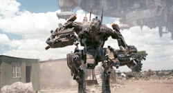 A large, dark, complex mechanical robot stands in a dusty outdoor setting under a cloudy sky.