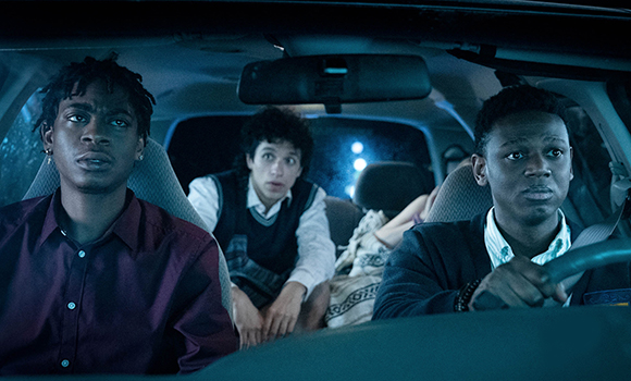 Three young men sit inside a car at night with expressions of shock and concern.