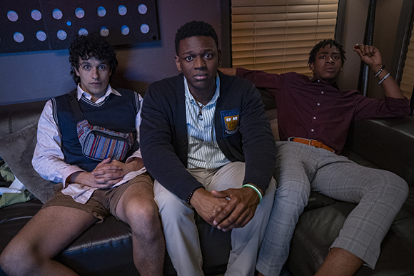 Three young men sit on a dark leather couch, looking toward the camera with various expressions.