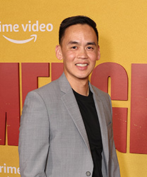 A smiling man wearing a grey blazer and black t-shirt in front of a yellow Prime Video backdrop.