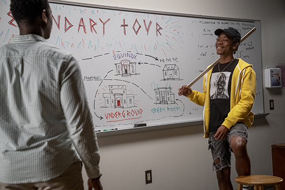 A person in a yellow hoodie points a stick at a whiteboard filled with drawings and text.