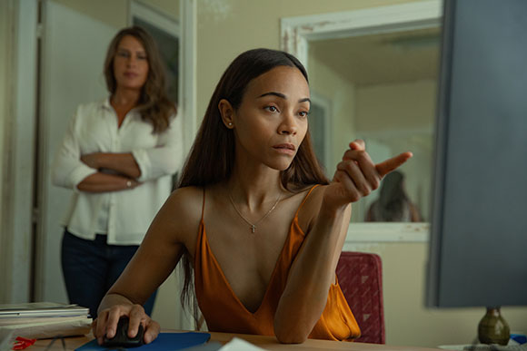 A woman in an orange top points at a computer monitor while another woman stands behind her with arms crossed.