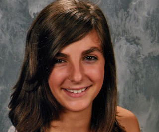 A young woman with long brown hair smiling against a mottled gray background.