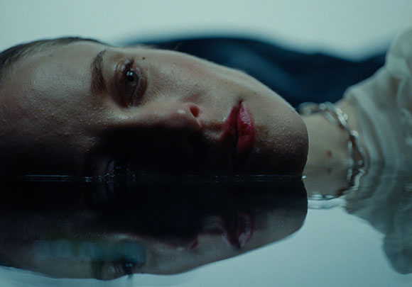 A close-up of a person's face floating in dark, still water with a reflection visible below.