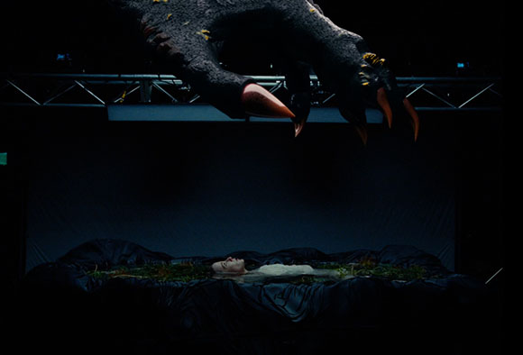 A large, dark, clawed creature hangs over a person lying on a dark, textured bed in a dark space.