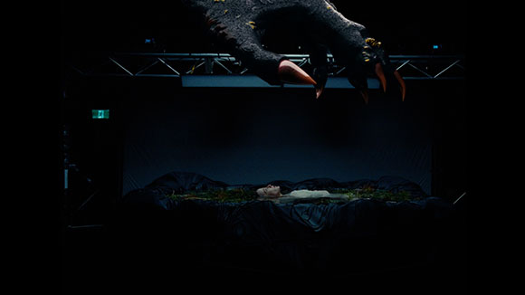 A massive, dark claw with sharp red tips hangs over a person lying on a dark, textured surface.