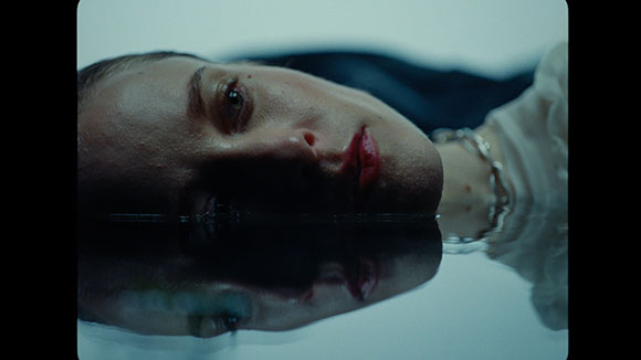A close-up of a person's face resting on the surface of dark, still water with a reflection below.