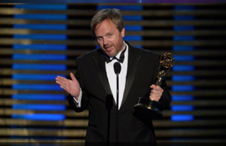 A man in a black tuxedo and bowtie holds a gold trophy while speaking at a podium.