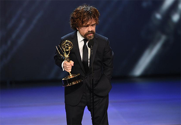 A man with curly hair and a beard in a dark suit holds a gold Emmy award while standing at a microphone.