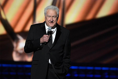 An older man with white hair wearing a black suit and tie, speaking into a microphone on a stage.