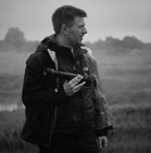 A man in a dark jacket holding binoculars while looking off to the side in a misty field.