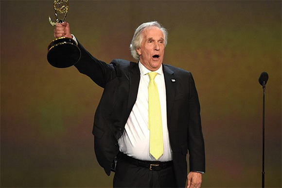 A man in a black suit and yellow tie holds a gold trophy aloft with an excited expression.