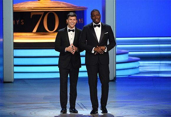 Two men in black tuxedos and bowties standing on a blue stage in front of a large gold number 70.