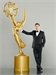 A man in a black tuxedo stands beside a giant gold Emmy Award statue against a gray background.