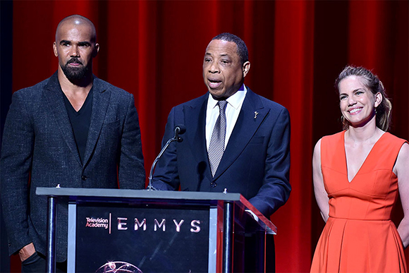 Three people stand behind a podium with the Emmy Awards logo against a red curtain.