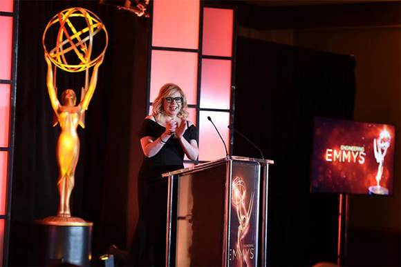 A woman in black stands at a podium next to a large gold Emmy statue on a stage.