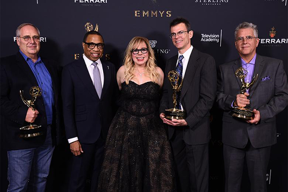 Five people in formal attire pose together, four of them holding gold Emmy awards against a dark backdrop.
