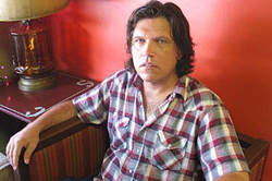 A man with medium-length dark hair wearing a red and white plaid shirt sits on a couch against a red wall.