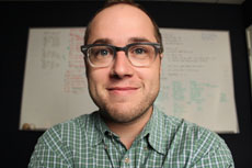 A man with glasses and a green checkered shirt smiles in front of a whiteboard with writing on it.