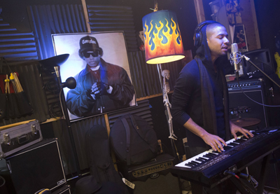 A man wearing headphones plays a digital keyboard in a studio filled with music equipment.