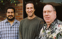 Three men smiling while standing in front of a brick wall and a dark window.