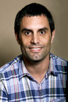 A man with short dark hair and a plaid shirt smiling against a neutral background.